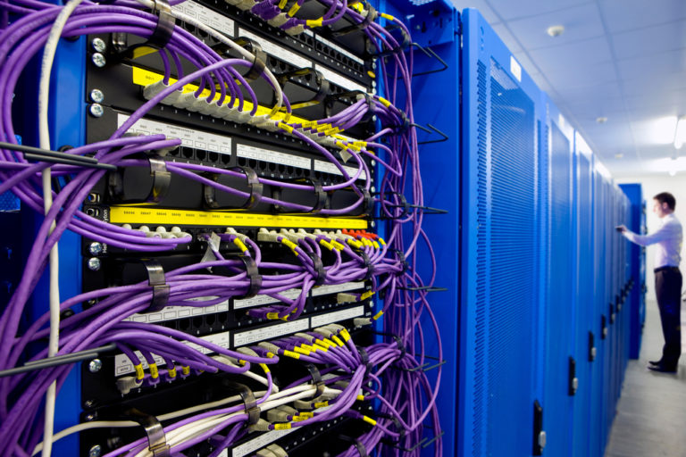 A close up shot of LAN cables and blue network servers with a man standing in background.