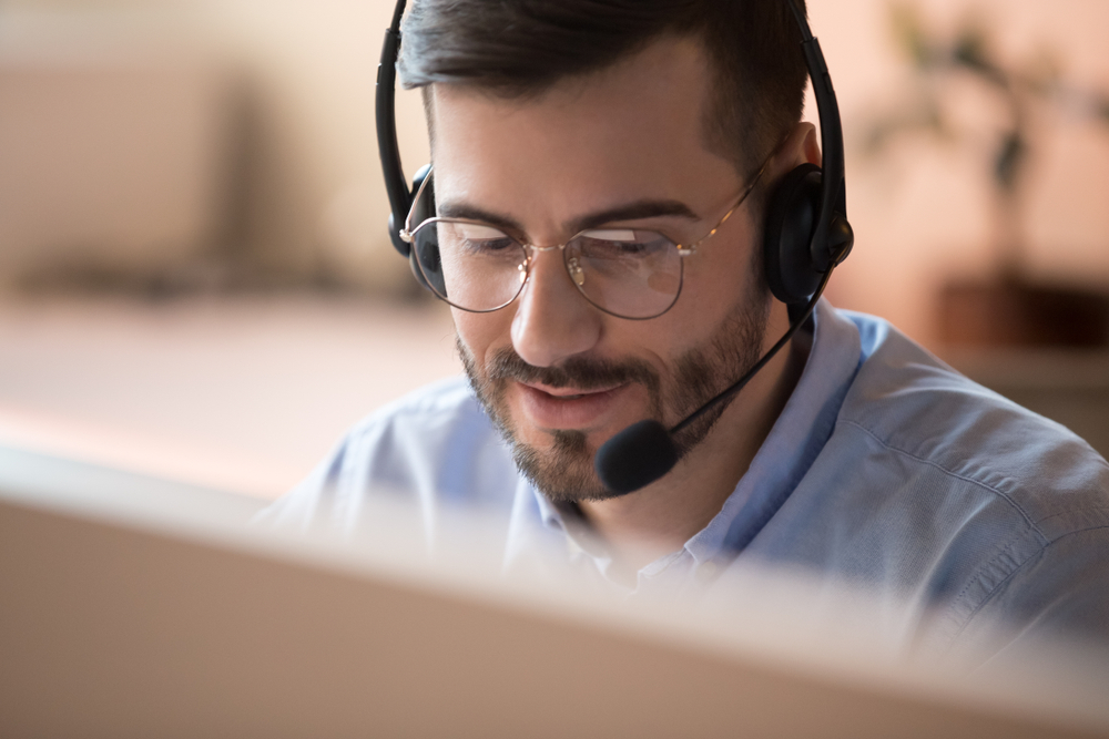 Focused IT support agent wear wireless headset make conference video call talk consult online client on computer, male helpline operator work in customer care support office